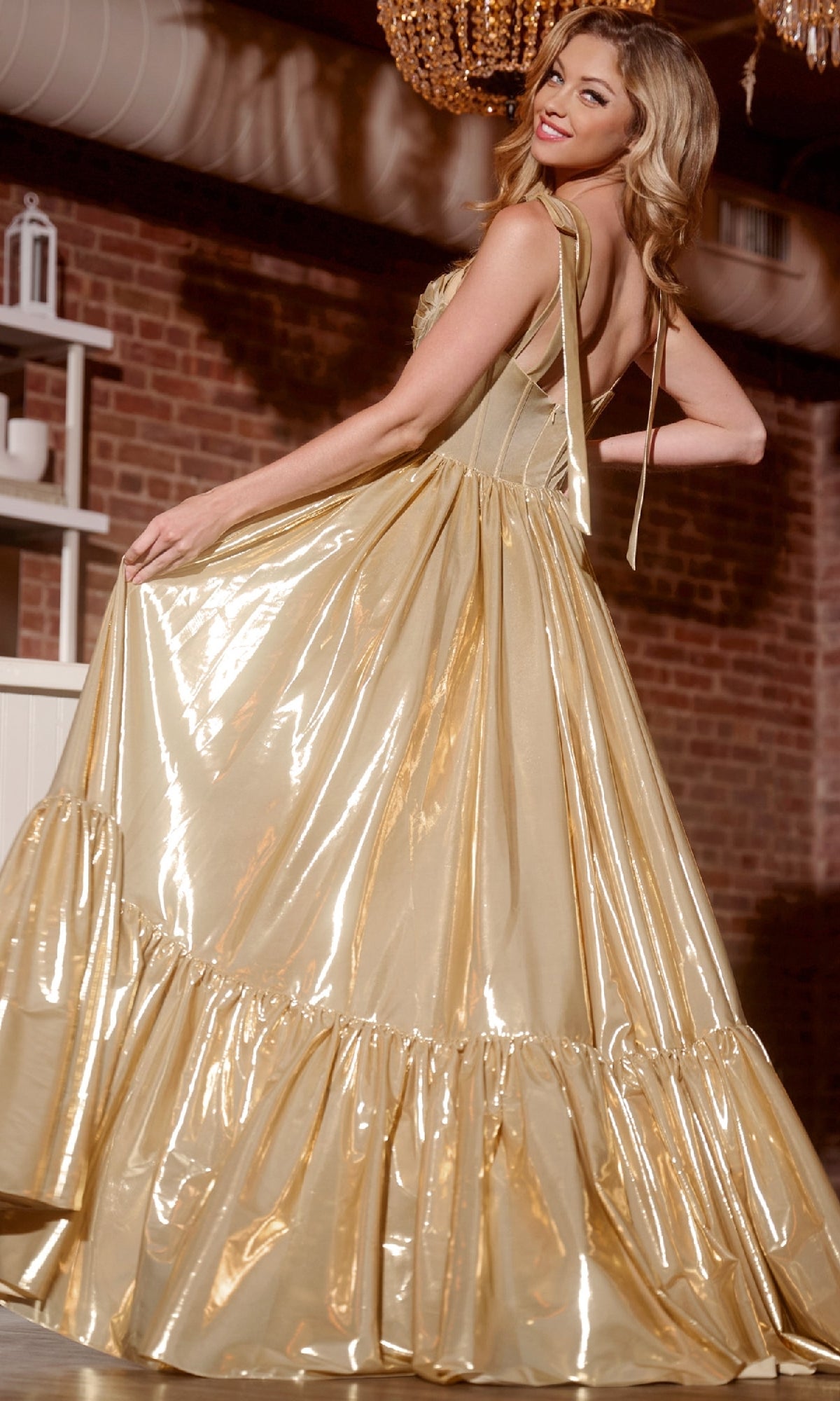 A woman poses indoors in front of a brick wall, looking over her shoulder and holding the skirt of the shiny gold Jovani 43596 long prom dress, featuring a corset bodice and ruffled hem.